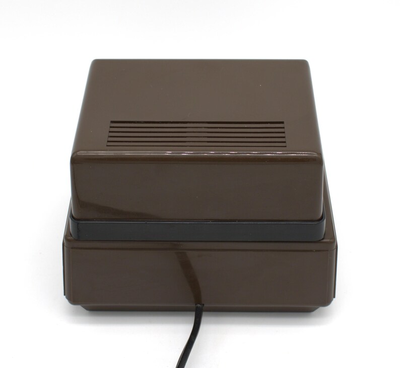 May include: A rectangular brown electronic device with a glossy finish and a black horizontal band separating the top and bottom sections. The top surface features a series of parallel ventilation slats, and a black power cord extends from the base.