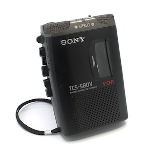 May include: Black Sony TCS-580V stereo cassette-corder with a built-in microphone and speaker. The device has a black wrist strap and the text "SONY" is visible on the front. The model number and "V-O-R" are also printed on the front.