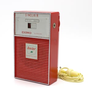May include: A red Sinclair Dino Supreme gas pump shaped transistor radio with a green Sinclair Gasoline logo and a yellow earbud cord.