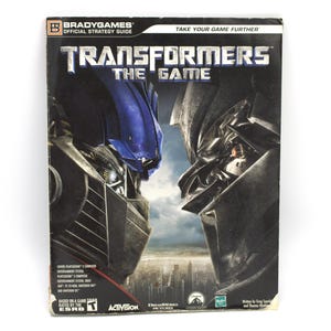 May include: A strategy guide for the video game "Transformers: The Game." The cover features a close-up of two robot faces, one blue and one gray, facing each other. The title is in large, silver letters. The BradyGames logo is in the upper left corner.