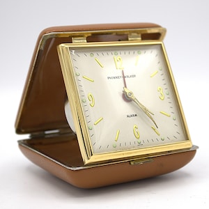 May include: A vintage brown leather travel alarm clock with a gold-tone case and a white face. The clock face has black numbers and hands, and the words "Phinney-Walker" and "Alarm" are printed in black. The clock is made in Japan.