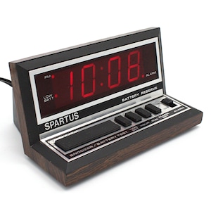 May include: A vintage Spartus digital alarm clock displaying 10:08 PM in red numerals. The clock has a black and wood-grain finish with buttons for snooze, time set, and alarm. The text "SPARTUS" is visible on the front.