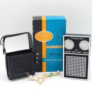 May include: A black and white portable transistor radio with a black carrying case and a white earbud. The radio has a blue and orange box with the text "Panorama De-Luxe Solid State Radio" on it.