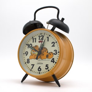 May include: A yellow twin-bell alarm clock with a cream-colored face featuring a graphic of the cartoon character Garfield. The clock has black metal bells, a top handle, and two pointed feet. The Sunbeam logo and Garfield text are visible.