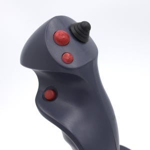 Puede incluir: Un joystick gris oscuro con tres botones rojos y una punta de goma negra. El joystick tiene un dise&ntilde;o curvo y probablemente se utiliza para jugar o controlar maquinaria.