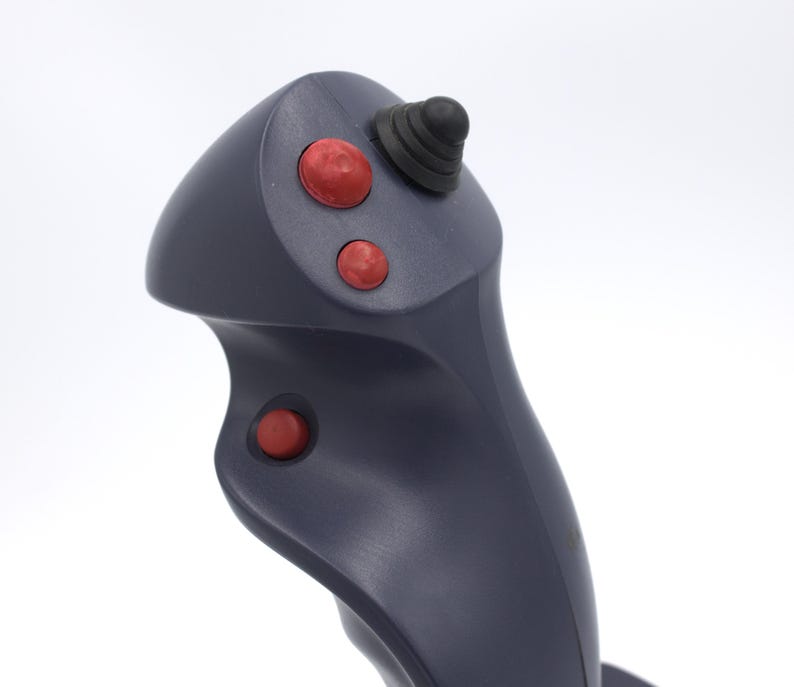 Puede incluir: Un joystick gris oscuro con tres botones rojos y una punta de goma negra. El joystick tiene un dise&ntilde;o curvo y probablemente se utiliza para jugar o controlar maquinaria.