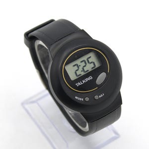 May include: Black digital talking watch with a round face. The watch displays the time 2:25 and has the word "TALKING" printed on the face. The watch has a black strap and buttons labelled "MODE" and "ADJ".