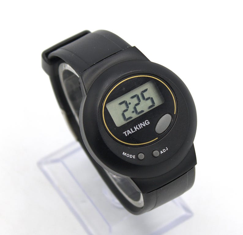 May include: Black digital talking watch with a round face. The watch displays the time 2:25 and has the word "TALKING" printed on the face. The watch has a black strap and buttons labelled "MODE" and "ADJ".