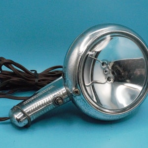 May include: A vintage chrome spotlight with a round reflector and a black cord. The spotlight has a switch on the handle.