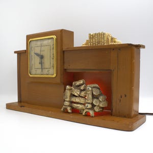 May include: A vintage brown wooden clock shaped like a fireplace. The clock has a gold-colored square face with black numbers and hands. The fireplace has a decorative log pile and a small light inside.