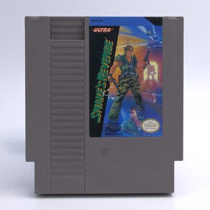 May include: A gray Nintendo Entertainment System (NES) game cartridge. The label features the title "Snake's Revenge" with a soldier illustration. The cartridge is marked "Made in Japan" and has the Ultra Games logo.