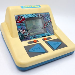May include: A yellow and blue handheld electronic game with the words "Space Alien" on the front. The game has a small screen and two buttons labeled "Left" and "Right".