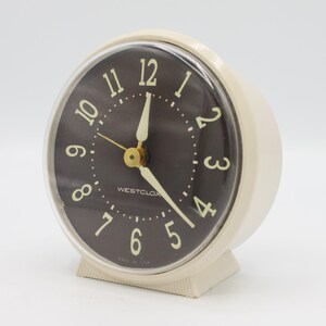 May include: A white vintage Westclox alarm clock with a black face and gold hands. The clock has a clear plastic cover over the face. The clock is made in the USA.