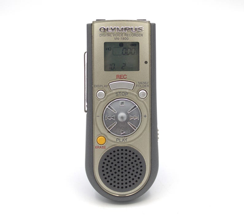 May include: A silver Olympus VN-1800 digital voice recorder with a black display screen, buttons, and a circular control knob. The screen displays "HO 000" and "10.2". The buttons include "REC", "STOP", "MENU/FOLDER", "DISPLAY", "PLAY", and "ERASE".