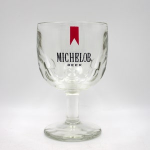 May include: A clear glass beer stein with a red and black Michelob Beer logo. The stein has a textured, dimpled surface.
