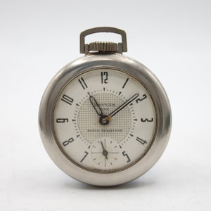 May include: A silver-toned pocket watch with a white face and black hands. The watch face features black numerals, a textured center, and the text "ONTDOX" and "SHOCK RESISTANT". A small seconds dial is at the bottom.