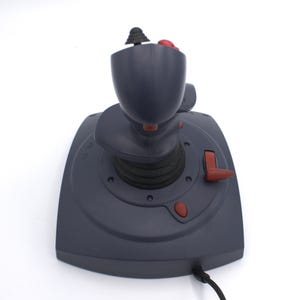 Puede incluir: Un joystick gris oscuro con una configuraci&oacute;n de botones negros y rojos. El joystick tiene una parte superior redondeada y una base con botones rojos adicionales. Un cable negro se extiende desde la base.