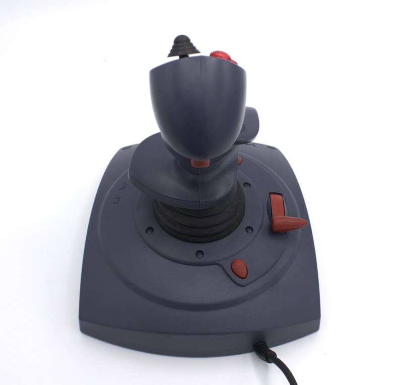 Puede incluir: Un joystick gris oscuro con una configuraci&oacute;n de botones negros y rojos. El joystick tiene una parte superior redondeada y una base con botones rojos adicionales. Un cable negro se extiende desde la base.
