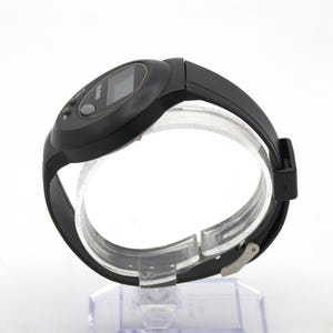May include: Black digital wristwatch with a black rubber strap. The watch face has a small rectangular display and two buttons. The word "TALKING" is printed on the watch face. The watch is displayed on a clear plastic stand.