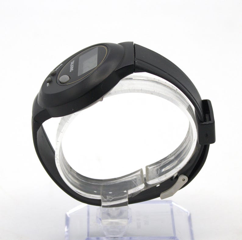 May include: Black digital wristwatch with a black rubber strap. The watch face has a small rectangular display and two buttons. The word "TALKING" is printed on the watch face. The watch is displayed on a clear plastic stand.