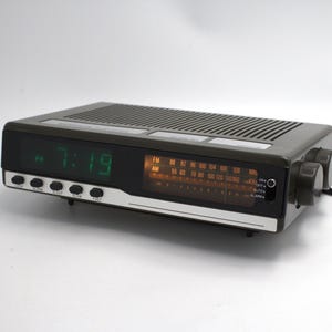 May include: A dark gray digital alarm clock with a green LED display showing 7:19. It has an FM/AM radio dial and control knobs on the side. The clock has several buttons and a silver trim.