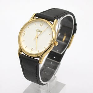 May include: A gold-toned wristwatch with a white face and black leather band. The watch face displays the word "TIME" and the numbers 3, 6, 9, and 12. The watch is displayed on a clear stand.