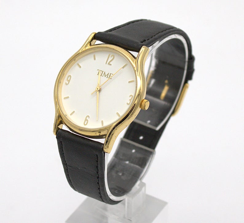 May include: A gold-toned wristwatch with a white face and black leather band. The watch face displays the word "TIME" and the numbers 3, 6, 9, and 12. The watch is displayed on a clear stand.
