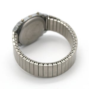 Peut inclure: Une montre num&eacute;rique argent&eacute;e avec un bracelet extensible. Le cadran de la montre est rond et le bracelet est compos&eacute; de petits maillons m&eacute;talliques rectangulaires. La montre a deux petits boutons sur le c&ocirc;t&eacute;.