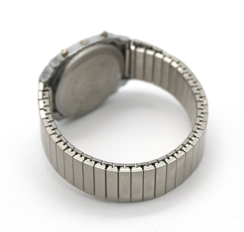 Peut inclure: Une montre num&eacute;rique argent&eacute;e avec un bracelet extensible. Le cadran de la montre est rond et le bracelet est compos&eacute; de petits maillons m&eacute;talliques rectangulaires. La montre a deux petits boutons sur le c&ocirc;t&eacute;.