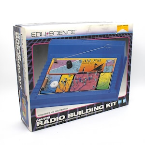 May include: An Edu Science AM/FM radio building kit in a blue box. The packaging features a graphic of the assembled blue radio console with colorful circuit board sections, electronic components, and a metal antenna. Text includes AM/FM RADIO KIT.