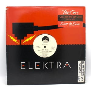 May include: A vinyl record album cover for The Cars' "You Are The Girl" from the album "Door to Door." The cover is black and red with the Elektra Records logo. The record label is white with black text.