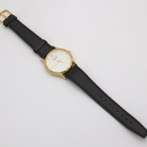 May include: A gold-toned wristwatch with a white face and black leather band. The watch face displays the word "TIME" and the number 12. The band is secured with a gold-colored buckle.