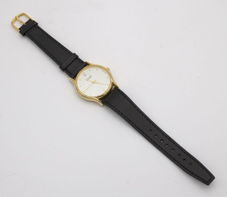 May include: A gold-toned wristwatch with a white face and black leather band. The watch face displays the word "TIME" and the number 12. The band is secured with a gold-colored buckle.