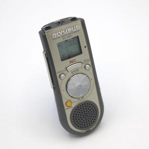 May include: A silver Olympus VN-1800 digital voice recorder with a black casing. The recorder has a small LCD screen, a record button, a play button, a stop button, a menu button, a folder button, and a display button. The recorder also has a built-in microphone and speaker.
