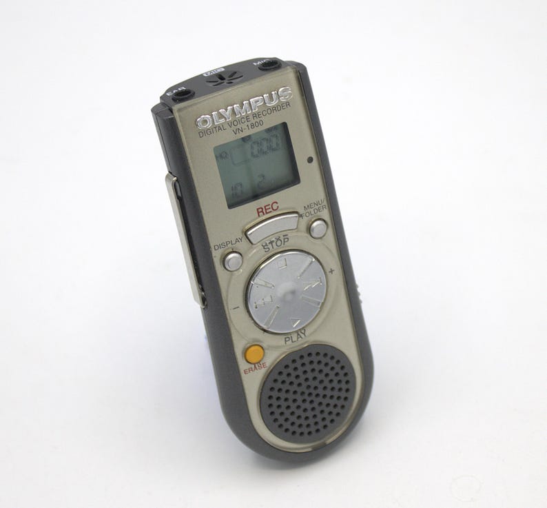 May include: A silver Olympus VN-1800 digital voice recorder with a black casing. The recorder has a small LCD screen, a record button, a play button, a stop button, a menu button, a folder button, and a display button. The recorder also has a built-in microphone and speaker.