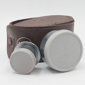 May include: Two gray lens caps with the text "SUN OPT." and a stylized "S" logo. The caps are sitting on a brown leather case.