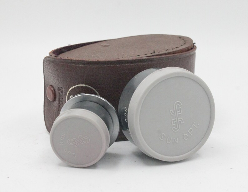 May include: Two gray lens caps with the text "SUN OPT." and a stylized "S" logo. The caps are sitting on a brown leather case.