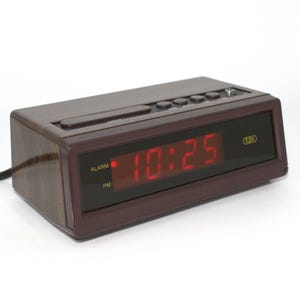 May include: A rectangular digital alarm clock with a dark brown faux wood grain finish. The front features a black display panel showing the red digital time 10:25, with illuminated text for ALARM and PM. Control buttons are located on the top surface.