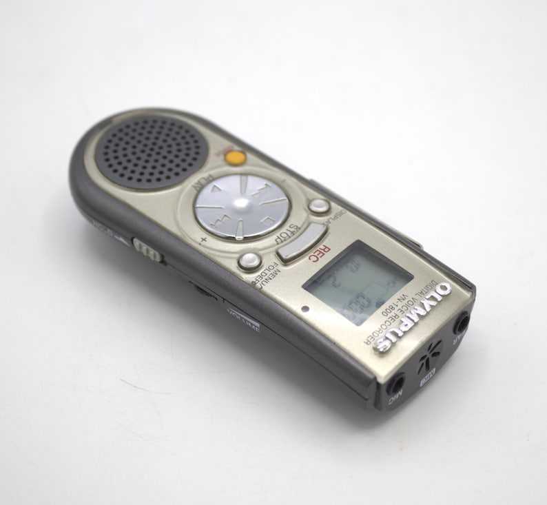 May include: A silver Olympus VN-1800 digital voice recorder with a black screen and buttons. The recorder has a built-in microphone and a headphone jack.