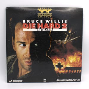 May include: A Laserdisc of the movie "Die Hard 2" featuring Bruce Willis. The cover art has a dark background with Bruce Willis's face on the left and a plane and fire on the right. The text "Die Hard 2" is in red.