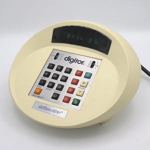 May include: Cream-colored Skillmaster calculator with a digital display showing "8 x 1 = 8." The calculator has a silver keypad with red, black, and white buttons. The word "digitor" is printed on the keypad.
