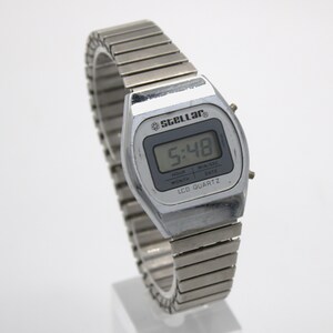 May include: A silver-toned digital wristwatch with a flexible metal expansion band. The watch face features the text Stellar, LCD Quartz, and displays the time 5:48. The rectangular screen includes labels for hour, month, min/sec, and date.