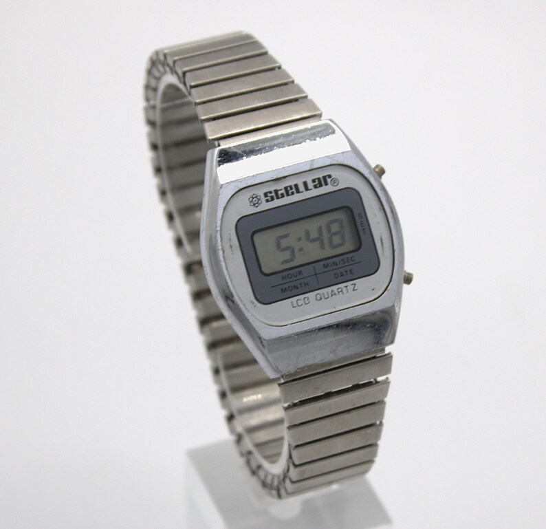 May include: A silver-toned digital wristwatch with a flexible metal expansion band. The watch face features the text Stellar, LCD Quartz, and displays the time 5:48. The rectangular screen includes labels for hour, month, min/sec, and date.