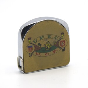 May include: A chrome and brass tape measure with a detailed emblem featuring a ship, flags, and text. The tape measure has a curved top and a metal belt clip. The emblem includes the letters "U.P.E.C." and "U.C.P."