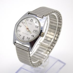 May include: Silver-toned Timex watch with a white face, featuring black numerals and hands. The watch has a date window at the 3 o'clock position and a silver expansion band. The watch face reads "TIMEX" and "WATER RESISTANT".