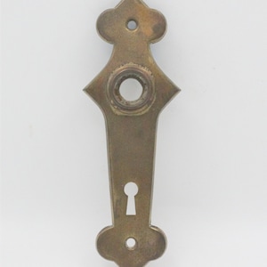 May include: A vintage brass door plate with a keyhole and decorative floral-like accents. The plate has a central circular opening and is approximately 7 inches long.