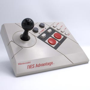May include: A light gray Nintendo NES Advantage joystick with a black joystick and red and gray buttons. The text "Nintendo NES Advantage" is printed in red on the front of the controller.