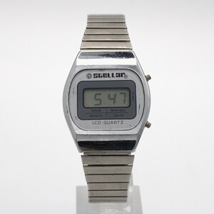 May include: A silver-toned digital wristwatch with a metallic link band. The watch face features the brand name Stellar, an atom icon, and the text LCD QUARTZ. The digital display shows the time 5:47, with labels for hour, min/sec, month, and date.