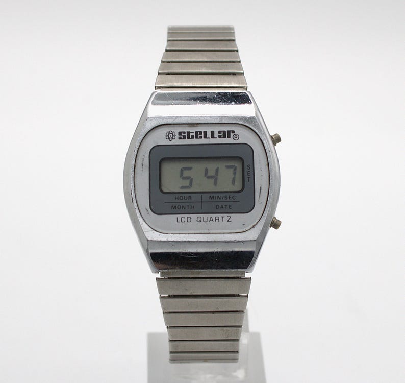 May include: A silver-toned digital wristwatch with a metallic link band. The watch face features the brand name Stellar, an atom icon, and the text LCD QUARTZ. The digital display shows the time 5:47, with labels for hour, min/sec, month, and date.