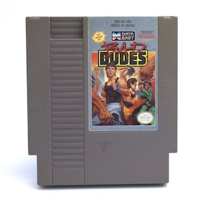 May include: A gray Nintendo Entertainment System (NES) game cartridge. The label features the game title "Bad Dudes" with action figures and the text "Top Arcade Hit!" and "Data East".